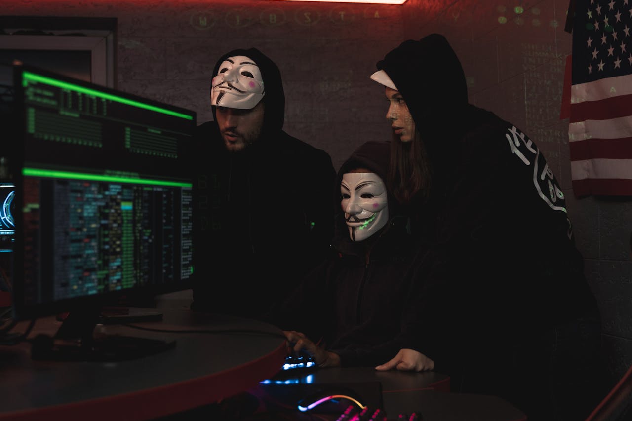 Masked hackers in hoodies working on a system in a dimly lit room, representing cyber security threats.