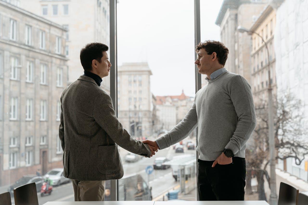 Two businessmen shaking hands in an urban office with city view through large windows.